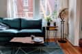 Living room with a teal sofa, wooden coffee table, globe, and geometric rug, overlooking a brick building through large windows.