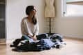 Woman sitting on floor with laundry pile, looking out window, dress form in background.