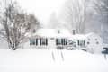 Snow-covered house with black shutters, surrounded by trees during a heavy snowfall.