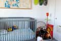 Child sitting on a pink stool in a nursery with a gray crib, colorful wall art, and skateboard decor.