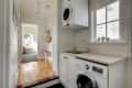 Laundry room with stacked washer and dryer, sink, and window with potted plants. Hallway leads to a sunlit room.