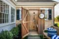 Outdoor shower with wooden doors, round mirror, and a bench with a blue striped bag, next to a house with green shutters.