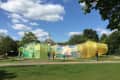 Colorful translucent pavilion in a park with people walking around, surrounded by trees and a historic building in the background.