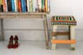 Rustic wooden table with books, red shoes underneath, and a colorful crocheted stool beside it.