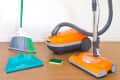 Orange vacuum cleaner with hose, green broom, teal dustpan, and sponge on wooden floor.
