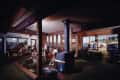 Cozy living room with people socializing, playing guitar, and dining by large windows overlooking a marina.