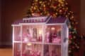 Pink dollhouse with multiple rooms, including a bedroom, living room, and bathroom, set in front of a decorated Christmas tree.