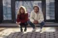 Two people crouching on a patterned rug in front of large windows, one holding a water bottle.