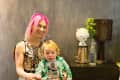 Woman with pink hair and child sitting on a chair, next to a table with a lamp and decorative items.