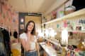 Woman in a dressing room with floral wallpaper, standing by a vanity filled with photos, plants, and colorful decor.