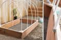 Wooden raised garden bed with white PVC hoop frame, set on gravel, next to a wooden fence and a shovel.