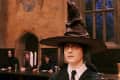 Young person wearing a large pointed hat in a medieval-style hall with candles and seated figures in the background.