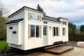 White tiny house on wheels with black roof, large windows, and a small wooden deck, set in a grassy area.