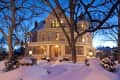 Victorian-style house with lit windows, surrounded by snow and trees, under a twilight sky.