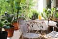 Small balcony with potted plants, white metal chairs, and a wooden table under a covered ceiling.
