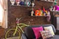 Exposed brick wall with colorful wigs on shelves, a green bicycle, and a dark sofa with vibrant pillows on a blue rug.