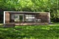 Modern tiny house with wooden siding, glass doors, and a patio set on a grassy lawn surrounded by trees.