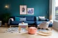 Modern living room with teal wall, blue sectional sofa, patterned cushions, pink pouf, and a beige rocking chair.