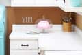 White kitchen with vintage blue fridge, pink cake on a stand, and "YUM" spelled on cork backsplash.