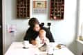 Man and baby sitting at a white table with mugs, books, and a baby bottle in a cozy room with wall shelves.