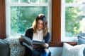 Woman reading a magazine on a window seat with cozy pillows and a view of greenery outside.