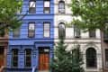 Blue townhouse with wooden door and steps, adjacent to a white townhouse, framed by green trees.