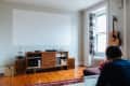 Living room with a wooden media console, turntable, guitar on wall, and person using a laptop on a brown sofa.