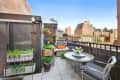 Rooftop patio with potted plants, a round table with flowers, and a cushioned bench with orange and black pillows.