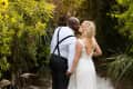 Bride in lace gown and groom in suspenders kiss on a gravel path surrounded by greenery.