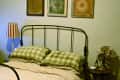 Bedroom with metal bed frame, checkered bedding, striped lamp, vintage fan, and three framed artworks on the wall.