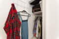 Small closet with hanging plaid shirt, denim overalls, and various clothes. Luggage stored on upper shelf.