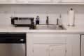 White kitchen cabinets above a stainless steel sink and dishwasher, with a paper towel holder and dish rack on the counter.