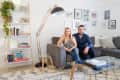 Couple sitting on a gray sectional sofa in a living room with a bookshelf, floor lamp, and framed photos on the wall.