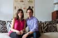 Couple sitting on a beige sofa with patterned cushions, under a seed dispersal poster, next to a white lamp.