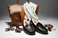 Brown leather tote with fur-trimmed gloves, cozy scarf, and black fur-lined slippers surrounded by pinecones.