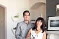 Couple smiling in living room holding a small white dog, with wall art and plants in the background.