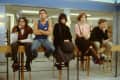 Five teenagers sitting on a library railing, each with distinct clothing styles, looking contemplative.