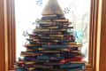 Stacked books forming a Christmas tree with red lights and a broom on top, set on a wooden windowsill.