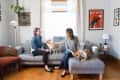 Two women sitting on a gray sofa with a dog and a goat in a cozy living room with framed art and plants.