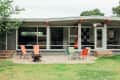 Mid-century modern house exterior with large windows, orange beams, and colorful patio chairs on gravel.