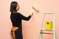 Woman painting a peach wall with a roller, standing next to a ladder with a yellow paint can.