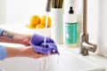 Hands washing a purple cloth under a kitchen faucet with dish soap, lemons, and lavender in the background.