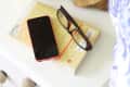 Nightstand with a gold lamp, book, red smartphone, glasses, and a wooden dish with a candle.