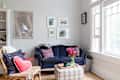 Cozy living room with a navy sofa, floral pillows, plaid ottoman, and wall art, next to large windows and a floor lamp.