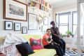 Couple sitting on a yellow sofa with a cat, surrounded by framed art, books, and plants in a bright living room.