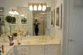 Bathroom with white vanity, gold fixtures, large mirror, and overhead lights. Person taking photo in reflection.
