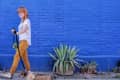 Woman walking a dog on a leash past a vibrant blue brick wall with potted plants.