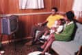 Family watching TV in a wood-paneled living room, sitting on a green sofa, child holding a doll.