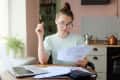 Woman with glasses reviewing documents at a kitchen table, with a laptop, calculator, and copper kettle in the background.