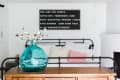 Bedroom with a metal bed frame, pink pillow, turquoise vase with branches, and a wall quote.
